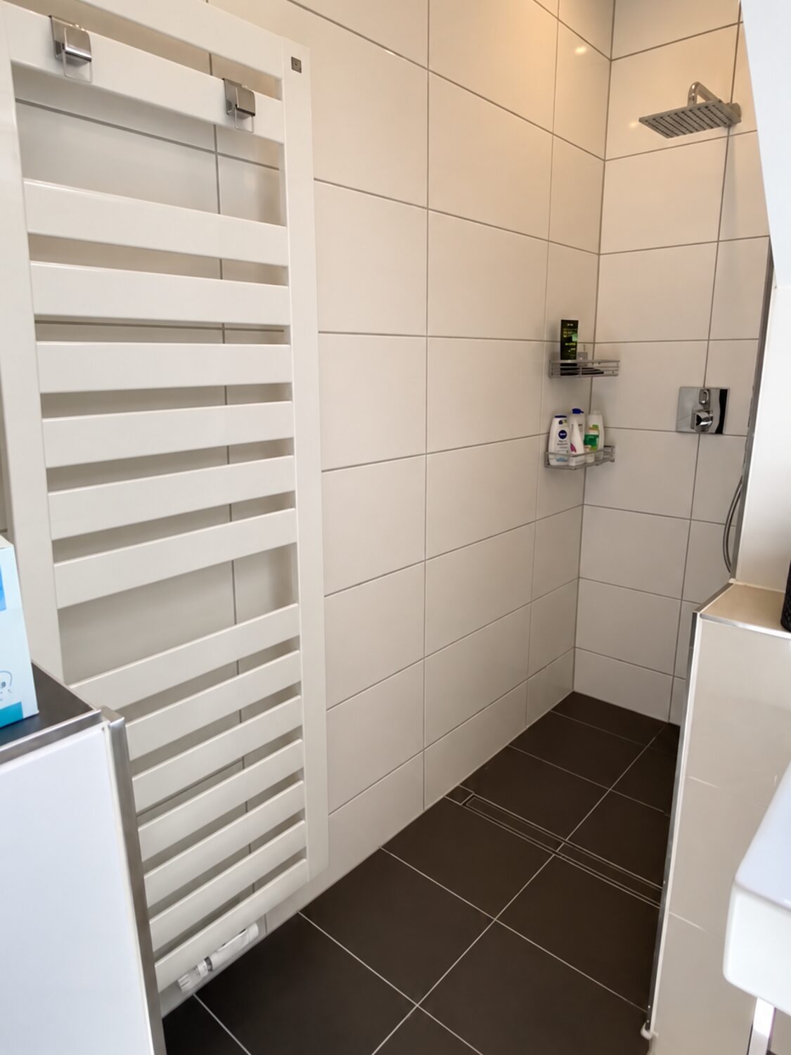 Die Dusche vorher: weiße Wandfliesen, dunkler Boden, weißer Handtuchheizkörper