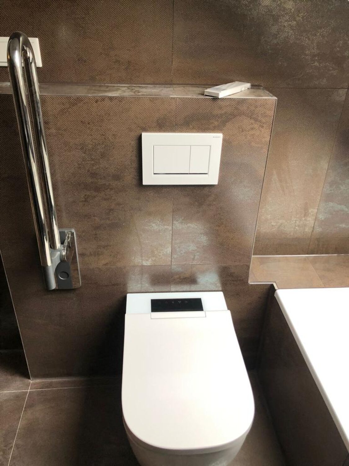 Dusch-WC mit Bedienfeld im Deckel vor einer Vorwand mit braunen Metalloptik-Fliesen, Kanten auf Gehrung geschnitten, klappbarer Stützgriff