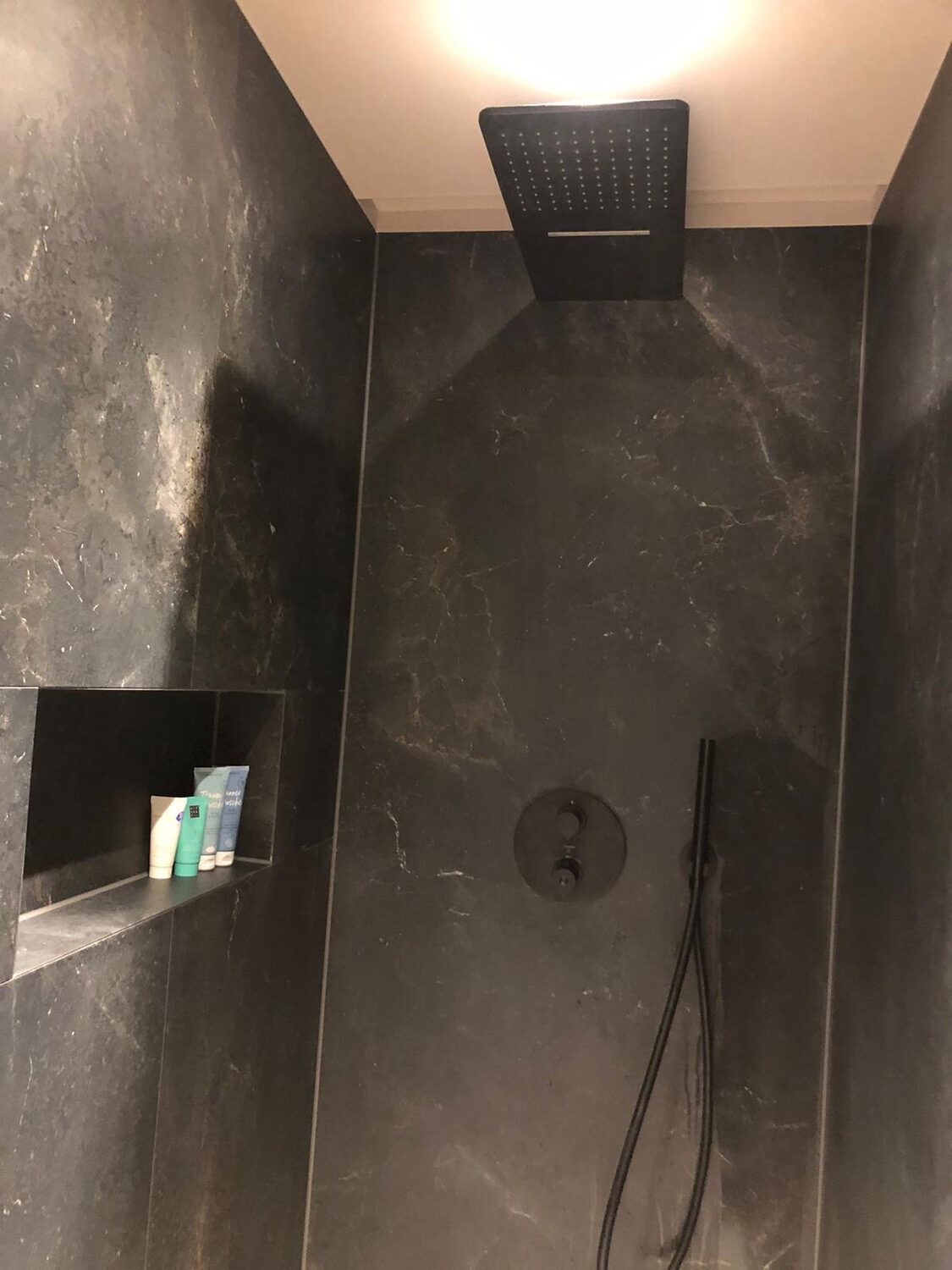 Dusche mit großformatigen Fliesen in dunkler Marmoroptik, schwarzer Deckenbrause und Wandnische