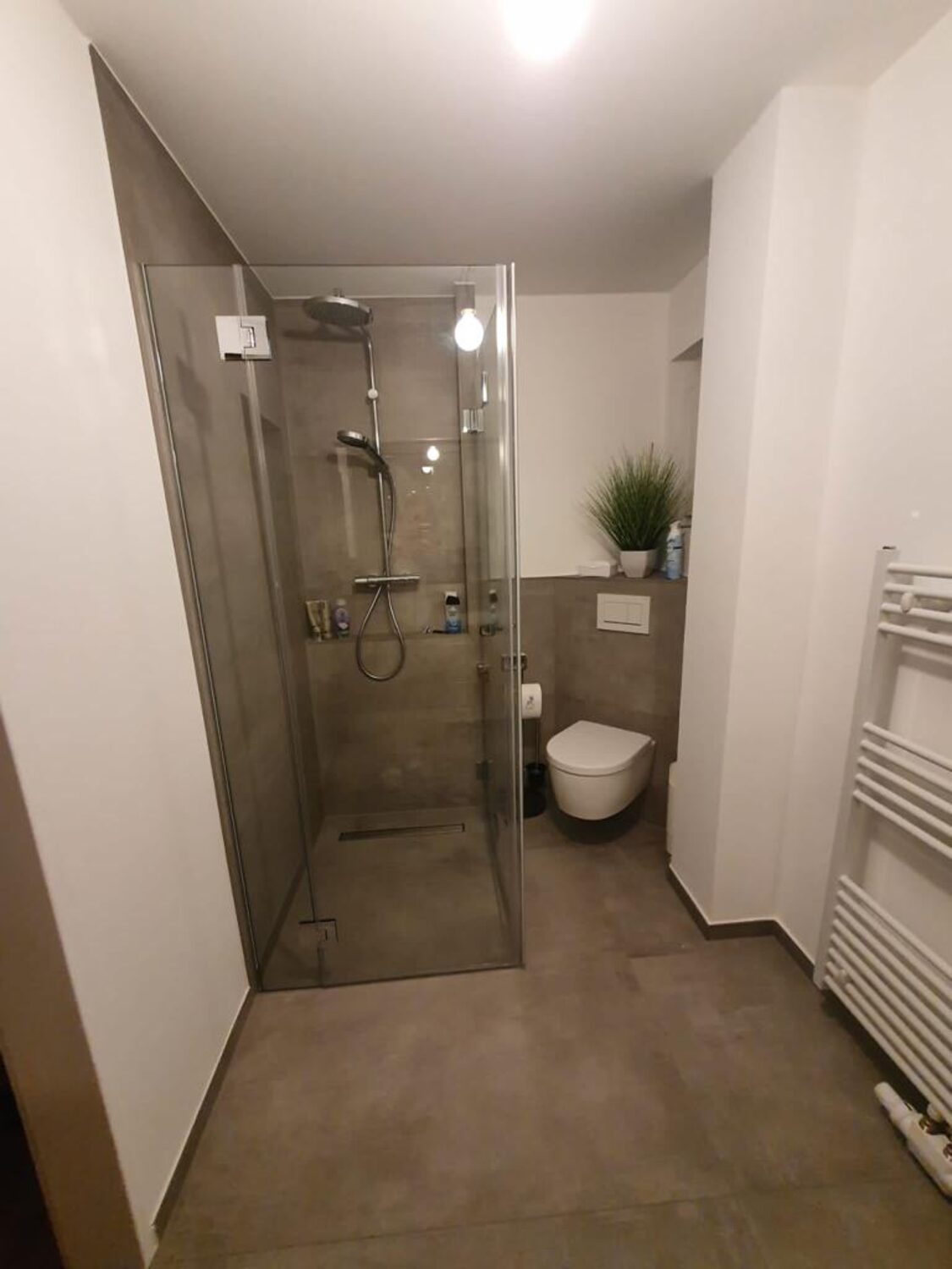 Bad mit gefliester Dusche in Betonoptik und gefliester WC-Vorwand, die übrigen Wände weiß verputzt, am Boden eine umlaufende Sockelfliese als Wischkante