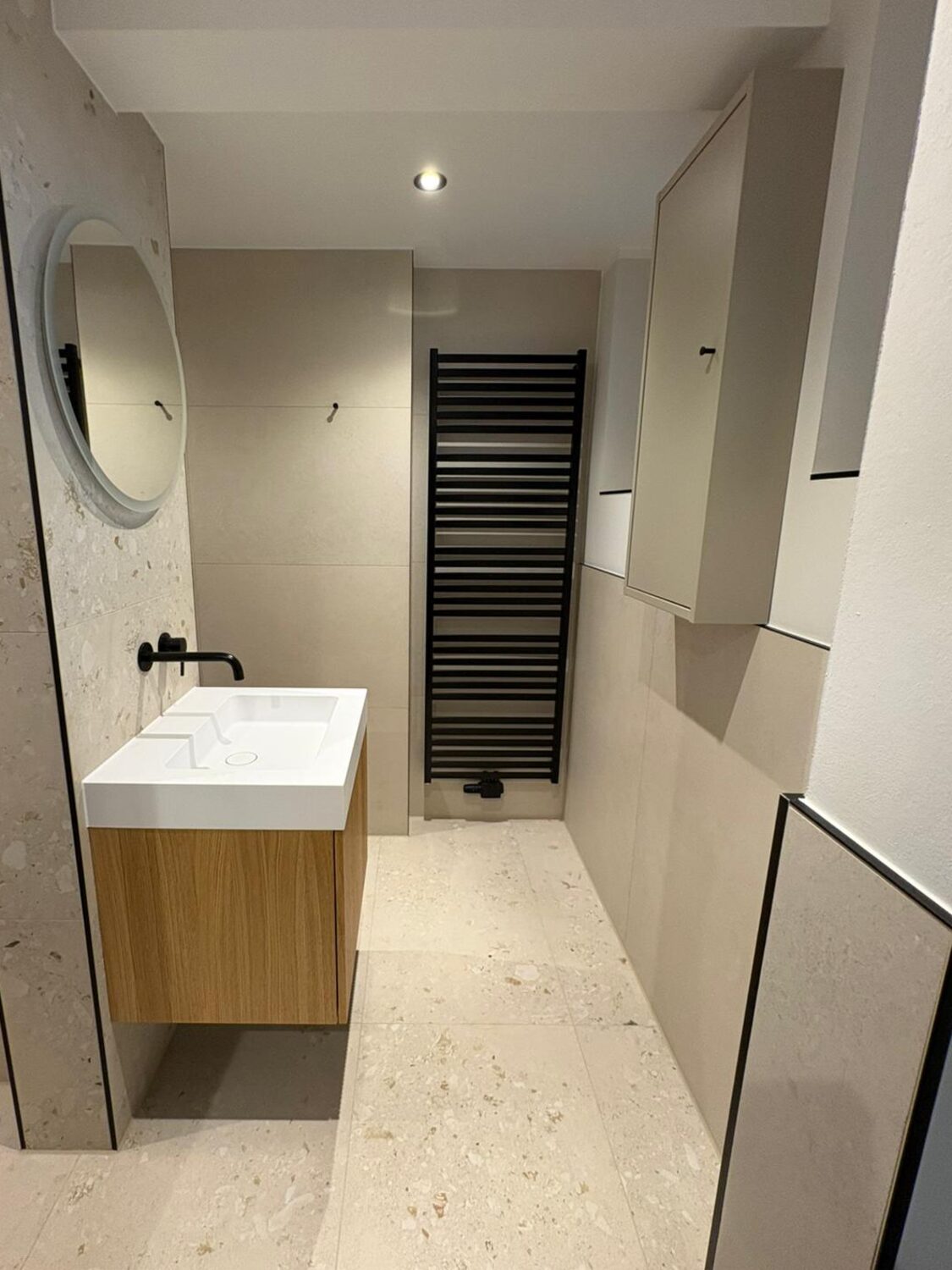 Bad mit beigen Terrazzo-Fliesen am Boden und an der Waschtischwand, Eichenunterschrank, schwarzer Wandarmatur und schwarzem Handtuchheizkörper